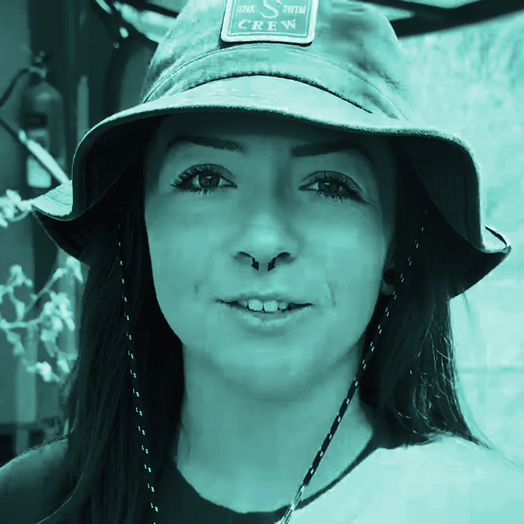 Lindsay Nikole, in a grey bucket hat, speaks to the lens outdoors in Kenya, beside an orange building and dry scrub.